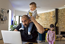 Ein Vater sitzt vor einem Laptop und trägt ein lachendes Kind auf den Schultern