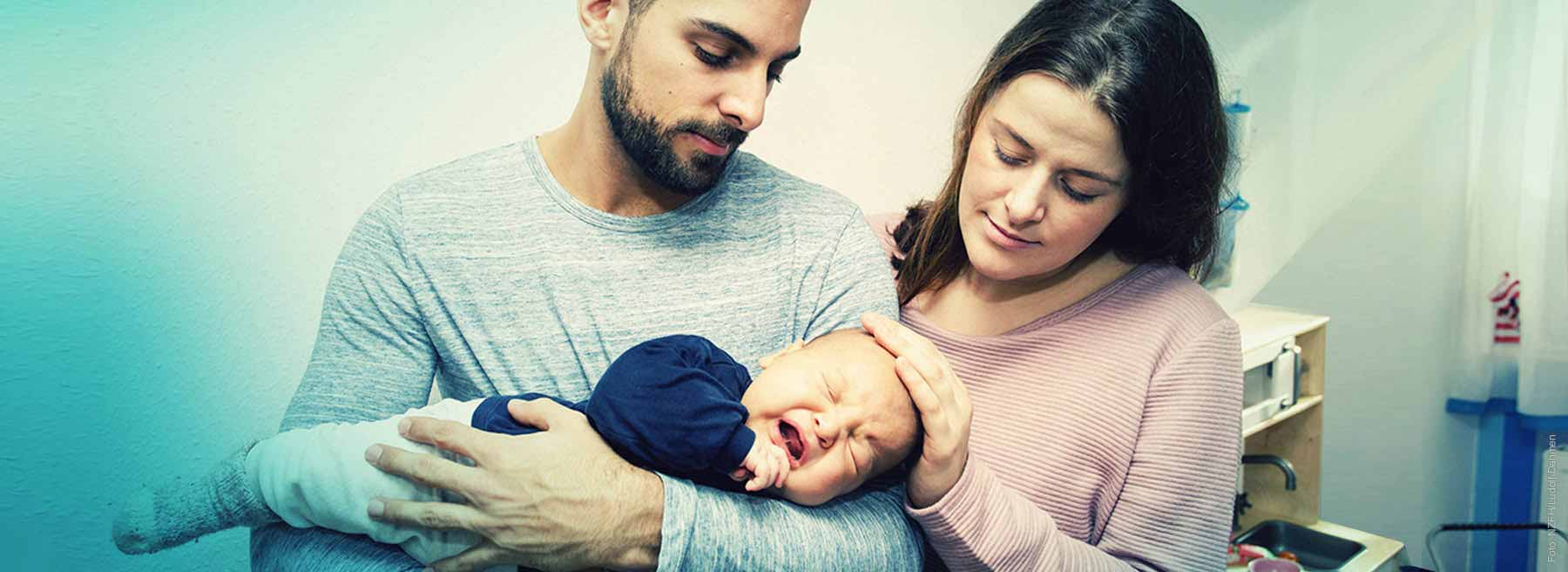 Mann mit schreiendem Baby auf dem Arm und Frau die daneben steht und ihre Hand auf das Köpfchen des Babys legt