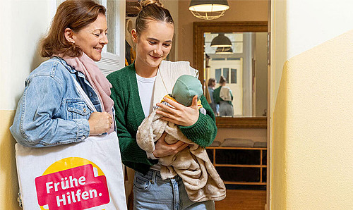 Junge Mutter begrüßt Besuchende der Frühen Hilfen an der Haustür und betrachtet mit ihr neutral das Baby