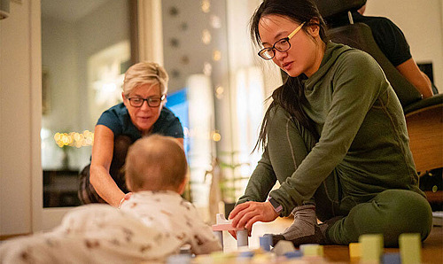 Mutter und Baby spielen mit Bauklötzen am Boden. Eine Familien-Gesundheits- und Kinderkrankenpflegende schaut zu.