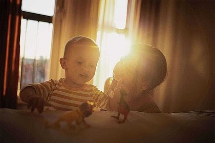 Eine Mutter spielt mit ihrem Kind in der Wohnung. Hinter Vorhängen scheint die Sonne.