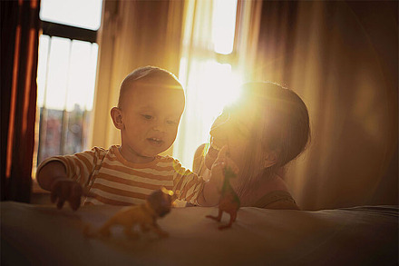 Eine Mutter spielt mit ihrem Kind in der Wohnung. Hinter Vorhängen scheint die Sonne.