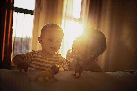 Eine Mutter spielt mit ihrem Kind in der Wohnung. Hinter Vorhängen scheint die Sonne.