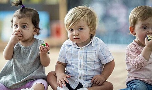 Drei Kleinkinder sitzen nebeneinander. Zwei von ihnen lutschen an Bauklötzen