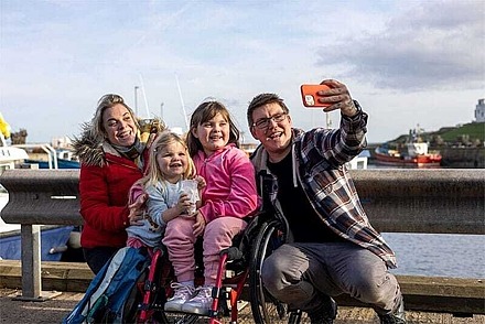 Eine Familie im Urlaub macht ein Selfie an einem Hafen. Eines der beiden Kinder sitzt im Rollstuhl.