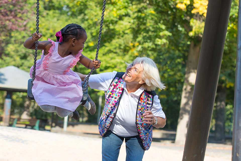 Familienpatin mit Kind auf dem Spielplatz Die Familienpatin schubst Kind auf der Schaukel an. Beide strahlen sich an.