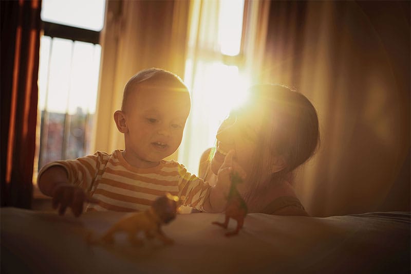 Eine Mutter spielt mit ihrem Kind in der Wohnung. Hinter Vorhängen scheint die Sonne.