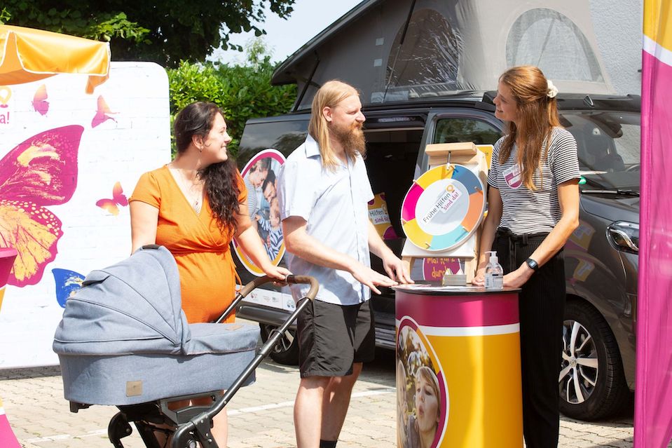 NZFH Mobile Fruehe Hilfen Vor dem Frühe-Hilfen-Bus steht ein Paar mit Kinderwagen und spricht mit einer Fachkraft