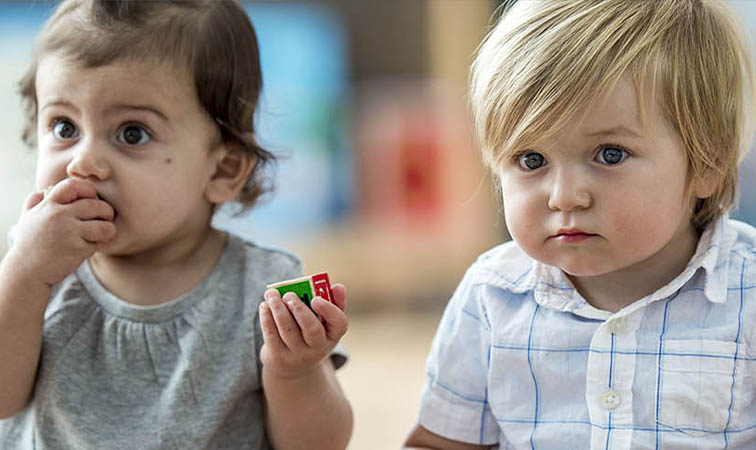 Zwei Kleinkinder sitzen nebeneinander. Das Mädchen lutscht an einem Bauklötzchen