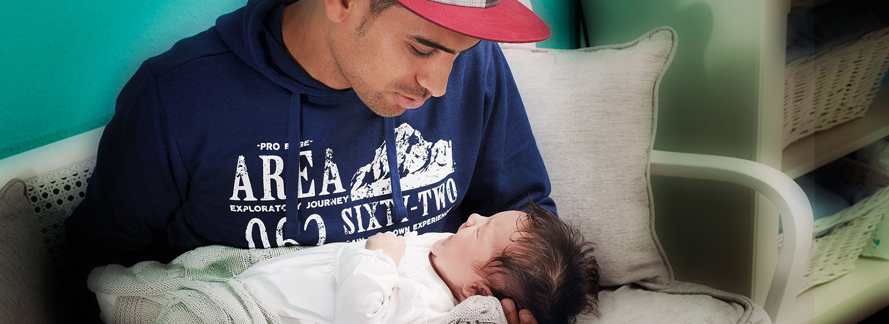 Vater mit Käppi und Baby auf dem Arm sitzt auf einem Sofa