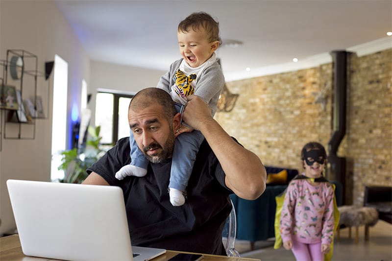 Ein Vater sitzt vor einem Laptop und trägt ein lachendes Kind auf den Schultern